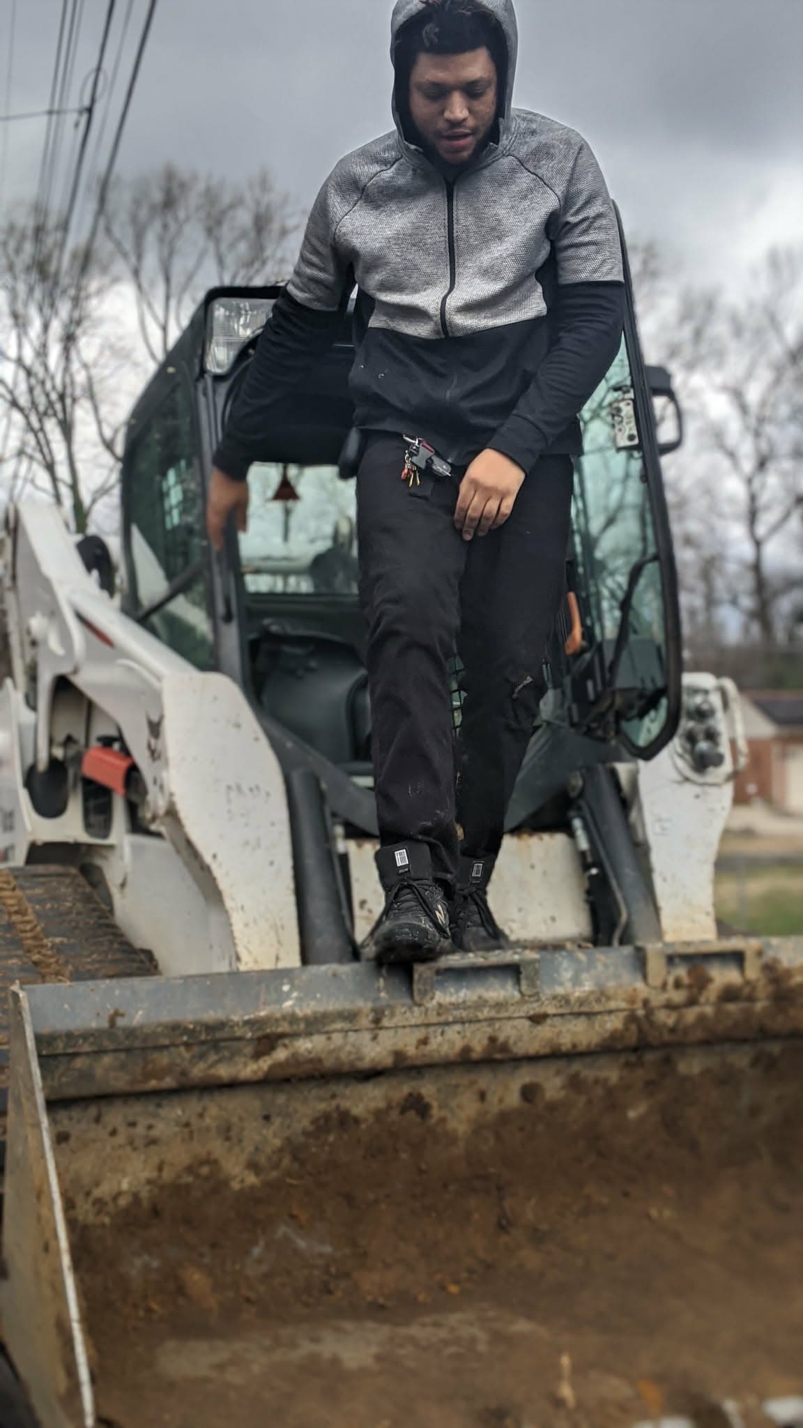 Man in a grey hoodie standing on the bucket of a white construction loader.