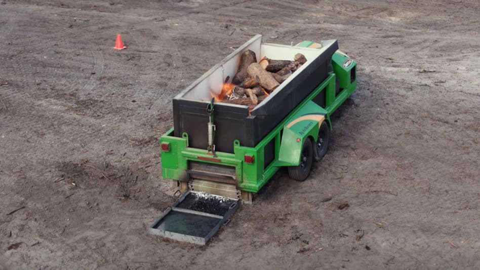 Green mobile burner incinerating wood logs and discharging biochar into a tray on dirt.